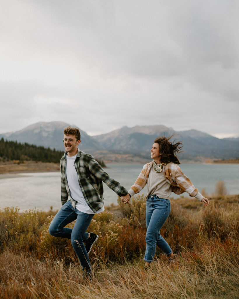 Denver Colorado Couple Session. Candid photo of a couple running through a grassy field with the Colorado landscape in the background.